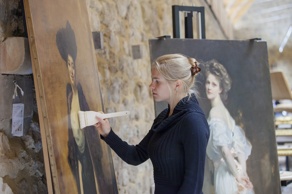 Paintings conservator brushing superficial dust from the surface of Duran's painting at Royal Oak Conservation Studio, Knole Barn, Kent. Nancy Witcher Langhorne, Viscountess Astor CH, MP (1879-1964) by John Singer Sargent and Margaret Anderson, The Hon. Mrs Ronald Greville DBE (1863-1942) by Emile-Auguste Carolus-Duran paintings side by side. Sargent after varnish removal, Duran before treatment